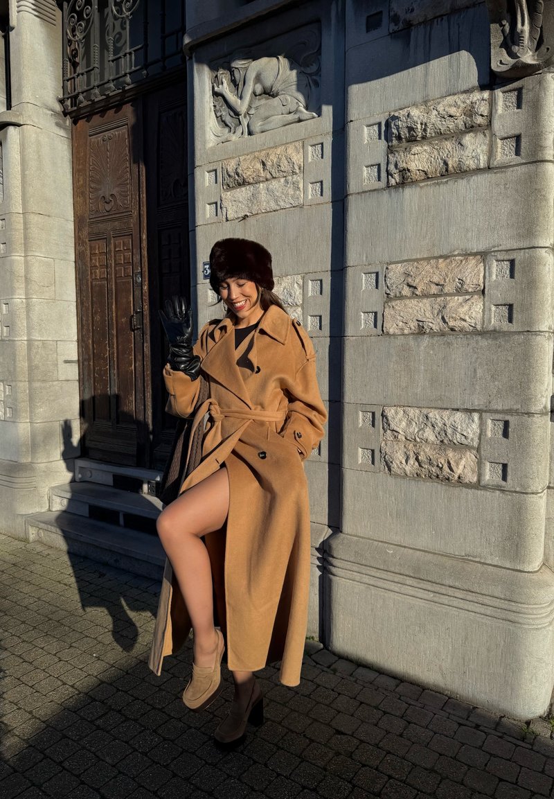 Trench-coat en laine beige avec ceinture, fente haute jusqu'à la cuisse, associé à des talons compensés marron et des gants en cuir noir. Un chapeau en fourrure complète le look.
