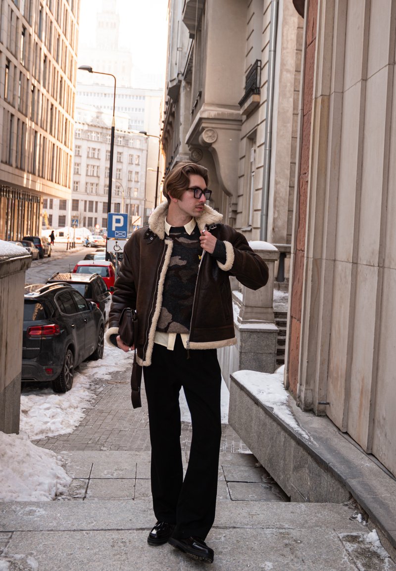 Homme en veste en peau lainée et lunettes marchant sur un trottoir enneigé en ville, à côté de voitures garées et de grands immeubles.
