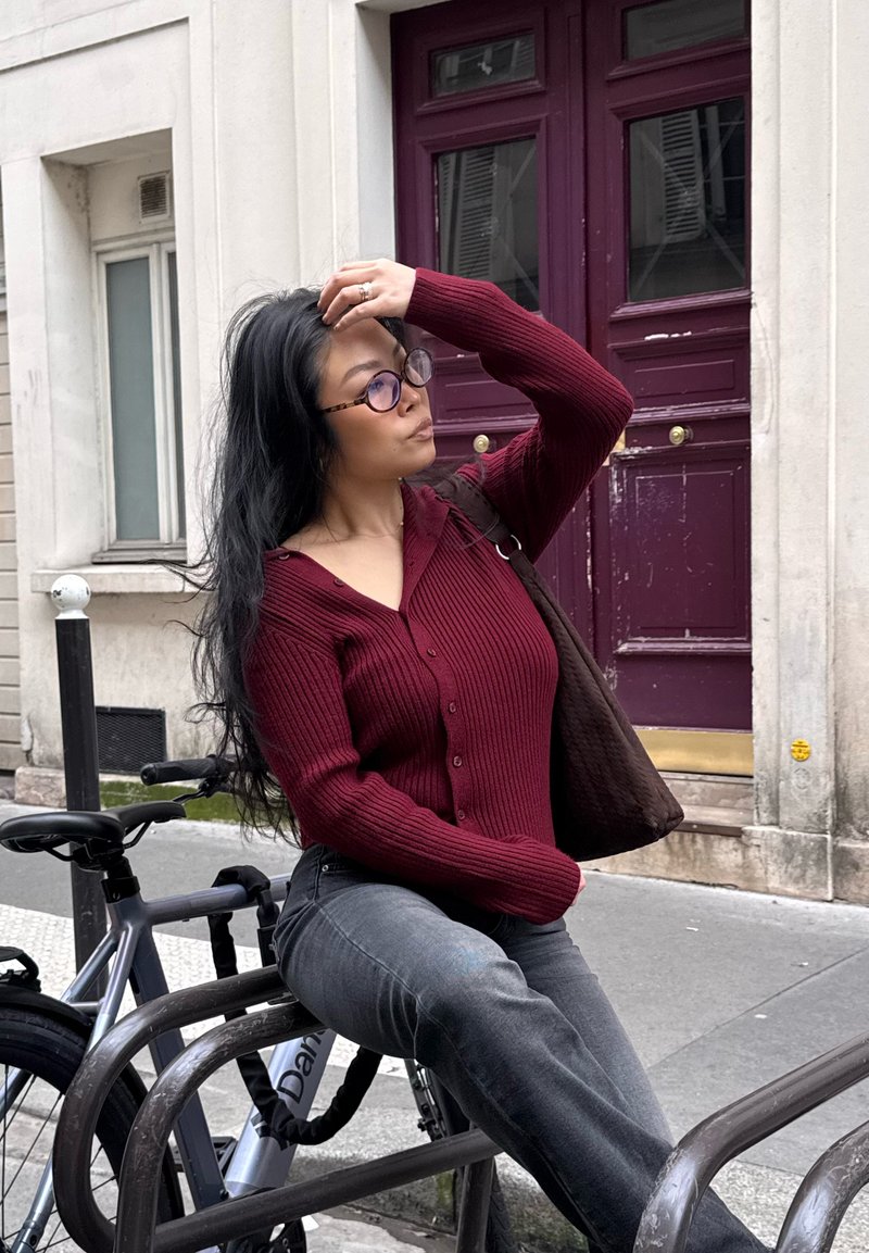 Femme en pull bordeaux et lunettes assise sur un support à vélos, touchant ses cheveux, avec un vélo attaché à proximité dans une rue de la ville.
