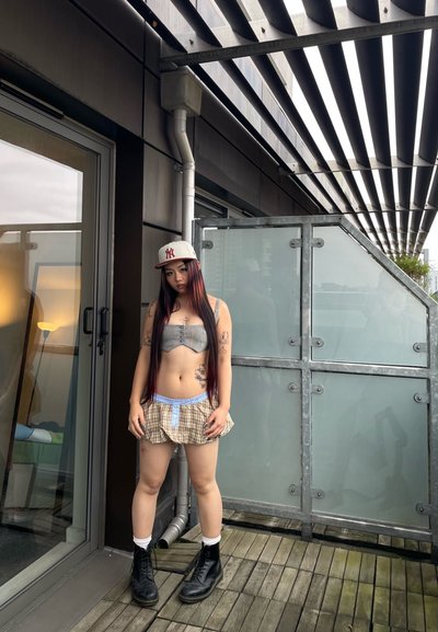 Una persona está de pie en una terraza exterior, vistiendo una camiseta corta de patrón y pantalones cortos de cuadros, combinados con botas negras y una gorra de béisbol.