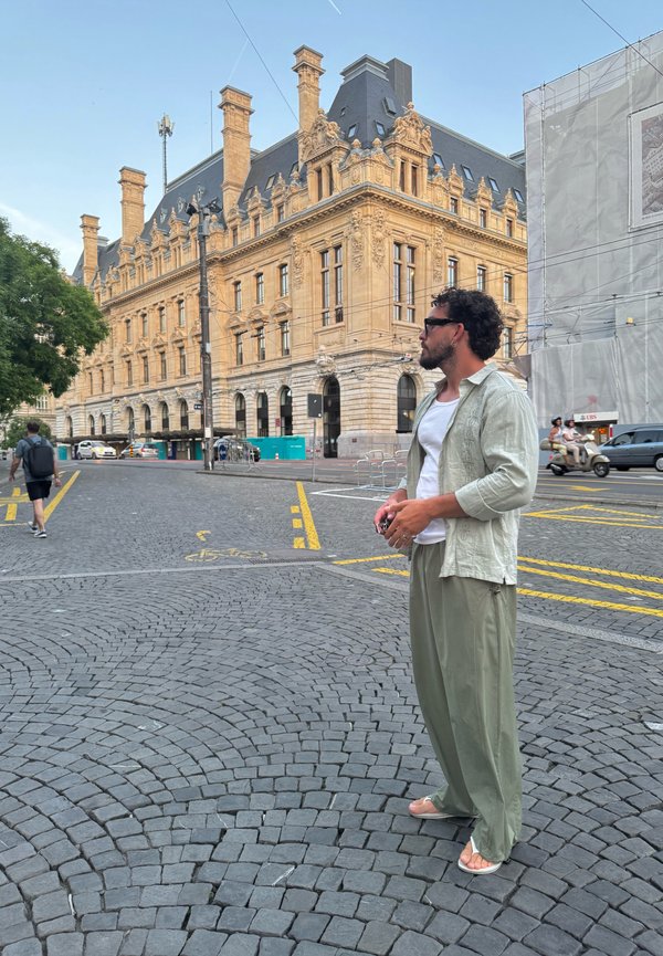 Hombre con gafas de sol y ropa verde claro está en una calle empedrada cerca de un edificio histórico ornamentado en un entorno urbano.