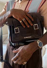 Personne portant des bagues et une montre tient une sacoche Dr. Martens marron foncé, vêtue d'un pull tricoté à rayures bleues et rouges.