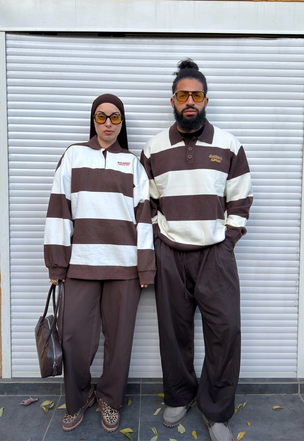 Kaksi henkilöä, jotka pukeutuvat samanlaisiin ruskeavalkoraidallisiin pitkähihnaisiin paitoihin ja ruskeisiin housuihin, seisovat valkoisen rullakaihtimen edessä, molemmilla keltaiset lasit.