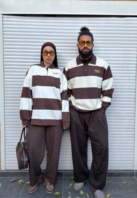 Kaksi henkilöä, jotka pukeutuvat samanlaisiin ruskeavalkoraidallisiin pitkähihnaisiin paitoihin ja ruskeisiin housuihin, seisovat valkoisen rullakaihtimen edessä, molemmilla keltaiset lasit.