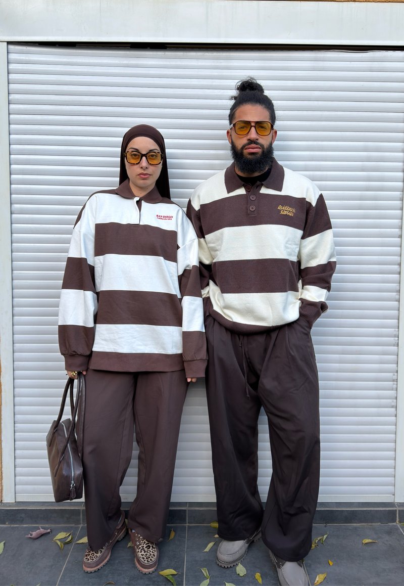 Zwei Personen, die identische braun-weiß gestreifte Langarmshirts und braune Hosen tragen, stehen vor einer weißen Jalousientür, beide mit gelb getönten Brillen.