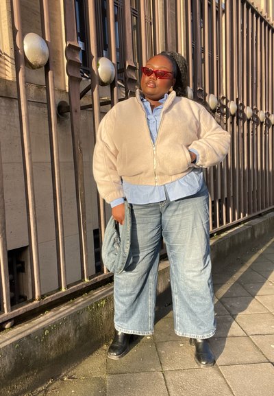 Chaqueta de forro polar marrón claro con cremallera sobre una camisa de botones azul claro, combinada con jeans de pierna ancha y botines negros. Gafas de sol rojas y un bolso azul.