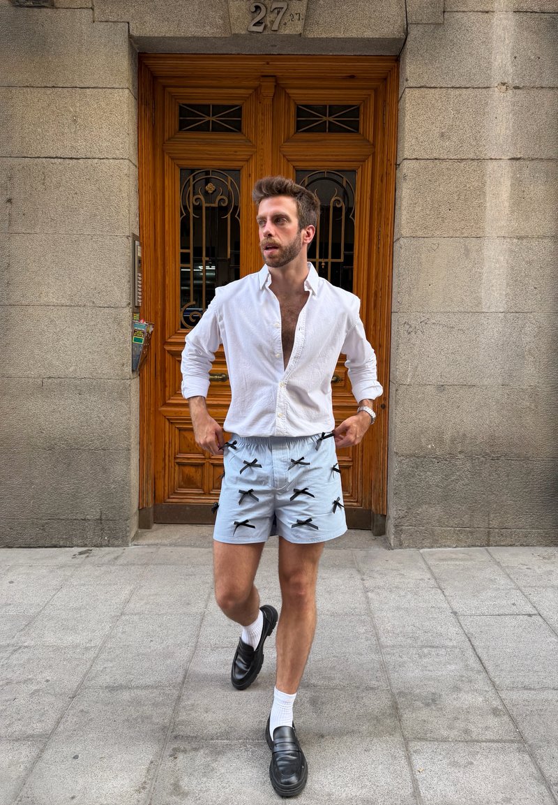 Homme portant une chemise blanche et un short clair avec des nœuds noirs, marchant sur le trottoir devant une porte en bois avec des grilles en fer.