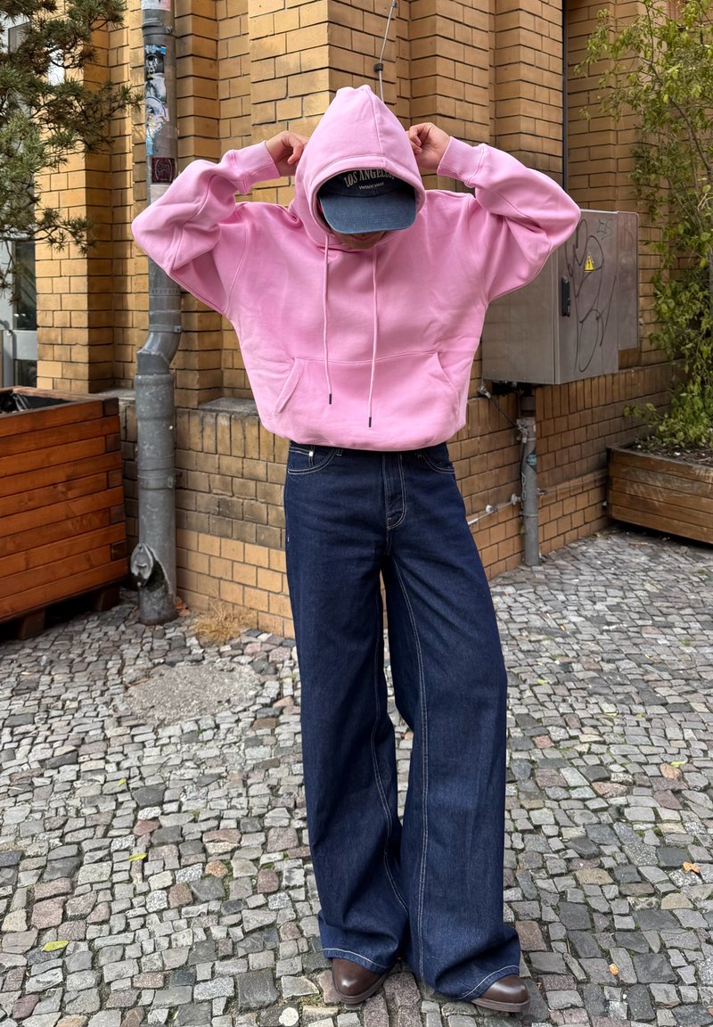 Pink hooded sweatshirt with adjustable drawstrings, paired with wide-leg navy jeans and brown shoes, set against a brick wall background.