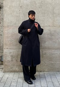 Fekete hosszú kabát magas gallérral, oldalsó zsebekkel és egyenes szabással, laza fekete nadrággal és fényes fekete cipővel párosítva, egy texturált fal előtt.