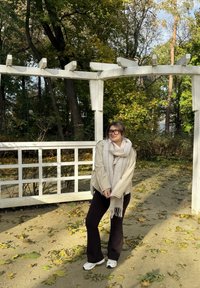 Beige yli-iso takki, kevyt huivi hapsuilla, mustat levenevät housut ja valkoiset tennarit. Maassa vihreitä ja keltaisia lehtiä.