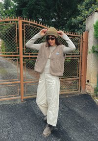 Brązowa kamizelka z polaru na jasnoszarym długim rękawie, kremowe szerokie spodnie i brązowe botki. Zieleń czapki i ciemne okulary słoneczne dopełniają całość.