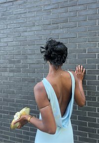 Svetlomodré saténové šaty s nízkym výstrihom a drapériou. Drží zlatú kruhovú kabelku s kvetinovými akcentmi. Pozadie z tmavej tehlovej steny.