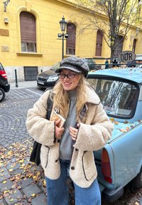 Beige flauschige Umpeljacke mit dunklem Besatz, grauer Pullover darunter, lockere blaue Jeans und eine Leopardenmuster-Mütze. Hält ein Buch in der Hand.
