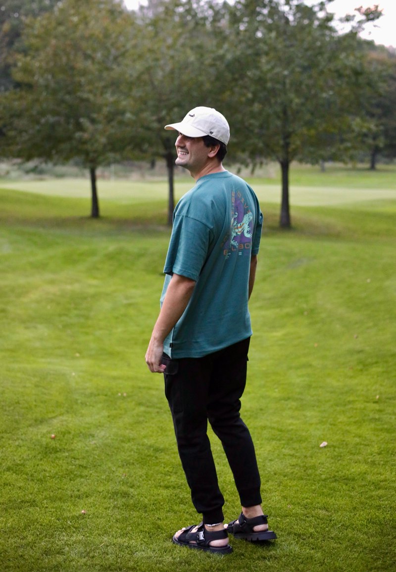 Tealfärgad oversized t-shirt med grafisk design, svarta joggers och svarta sandaler. Personen står i ett gräsområde med träd i bakgrunden.