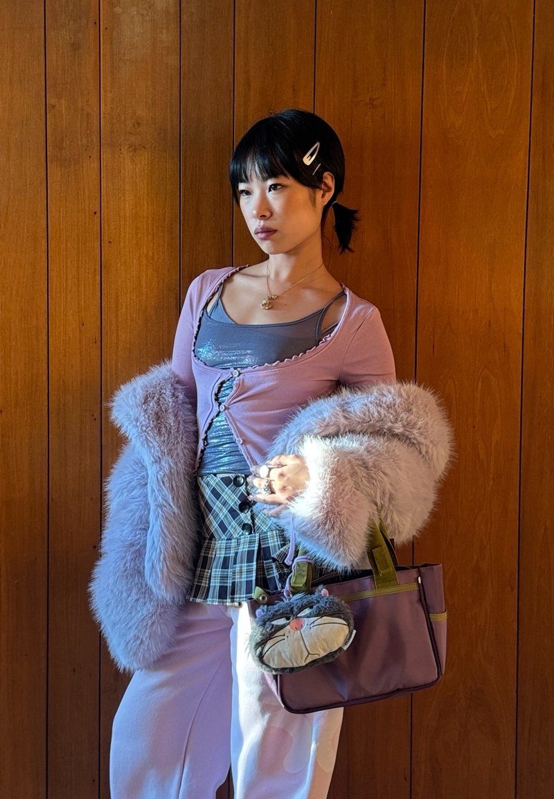 Jeune personne portant une veste duveteuse rose, une jupe à carreaux par-dessus un pantalon, tenant un sac violet avec un porte-clés en forme de visage de chat, debout devant un mur en bois.
