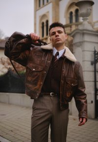 Brun skinnbomberjakke med en fôret krage i saueskinn, to brystlommer, brukt over en brun cardigan og stripet skjorte. Tilpassede grå bukser med tynne striper.