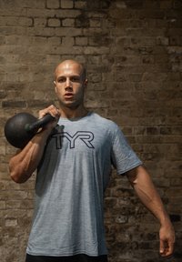 Graues Sport-T-Shirt mit dunkelblauem Logo, getragen von einer Person, die ein schwarzes Kettlebell vor einer strukturierten Steinwand hält. Deutliche Armdefinition sichtbar.