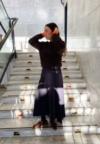 Maglione lavorato a maglia marrone, lunga gonna nera plissettata con dettagli in cintura, scarpe marroni, in piedi su scalini di marmo con luce solare e ombre.