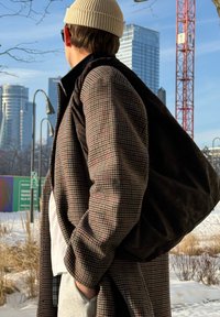 Homme portant un manteau à carreaux, un bonnet beige et des lunettes de soleil, transportant un sac noir en plein air dans une zone urbaine enneigée avec des bâtiments modernes et une grue de chantier.