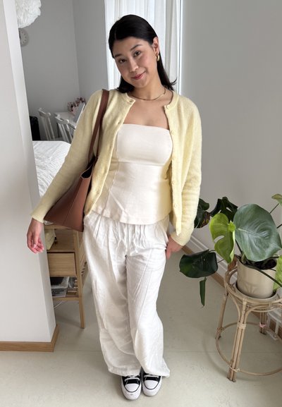 Cárdigan de punto amarillo claro, blusa blanca ajustada, pantalones sueltos de lino blancos y zapatillas negras. Bolso de hombro marrón y planta verde de fondo.