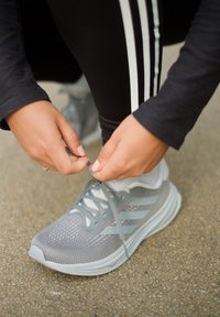 Grijze sportshoe met een bovenwerk van mesh, lichtgrijze accenten en een witte zool. Handen die de veters strikken, gekleed in zwarte legging met witte strepen.