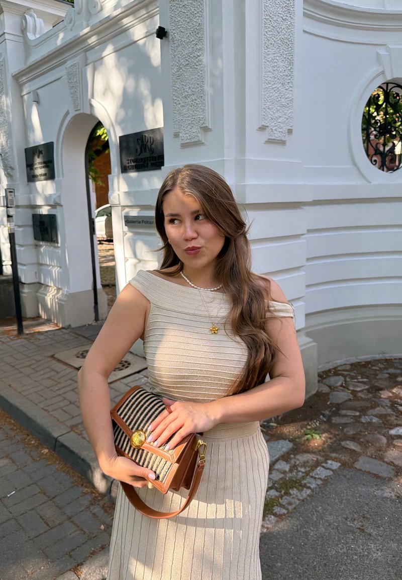 Jeune femme en robe beige épaules dénudées tenant un sac à main rayé se tient près d'un bâtiment blanc orné avec des plaques sur le mur.