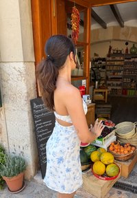 White sundress featuring a blue floral print and a cut-out design. Fresh fruits arranged in baskets, with wooden crates and a chalkboard menu in view.