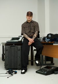 Košela s krátkym rukávom s károvaným vzorom, čierne nohavice, biele tenisky. Doplnky zahŕňajú hnedú čiapku, slúchadlá a elektronické zariadenia na stole.