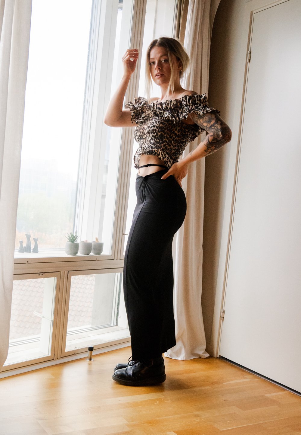 Top de leopardo com decote à americana, saia preta de cintura alta e sapatos pretos. Iluminação natural, piso de madeira e cortinas suaves visíveis.