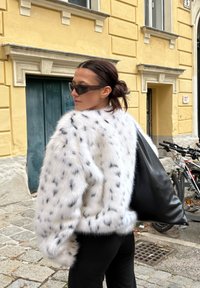 Bela jakna iz umetnega krzna s črnimi madeži, dolgimi rokavi in rebrastimi manšetami, kombinirana s črno usnjeno torbico in temnimi sončnimi očali.