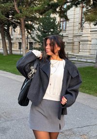 Giovane donna che indossa giacca grigia e gonna, maglione bianco, gioielli dorati e porta una borsa nera, in piedi vicino a degli alberi e a un edificio ornato.