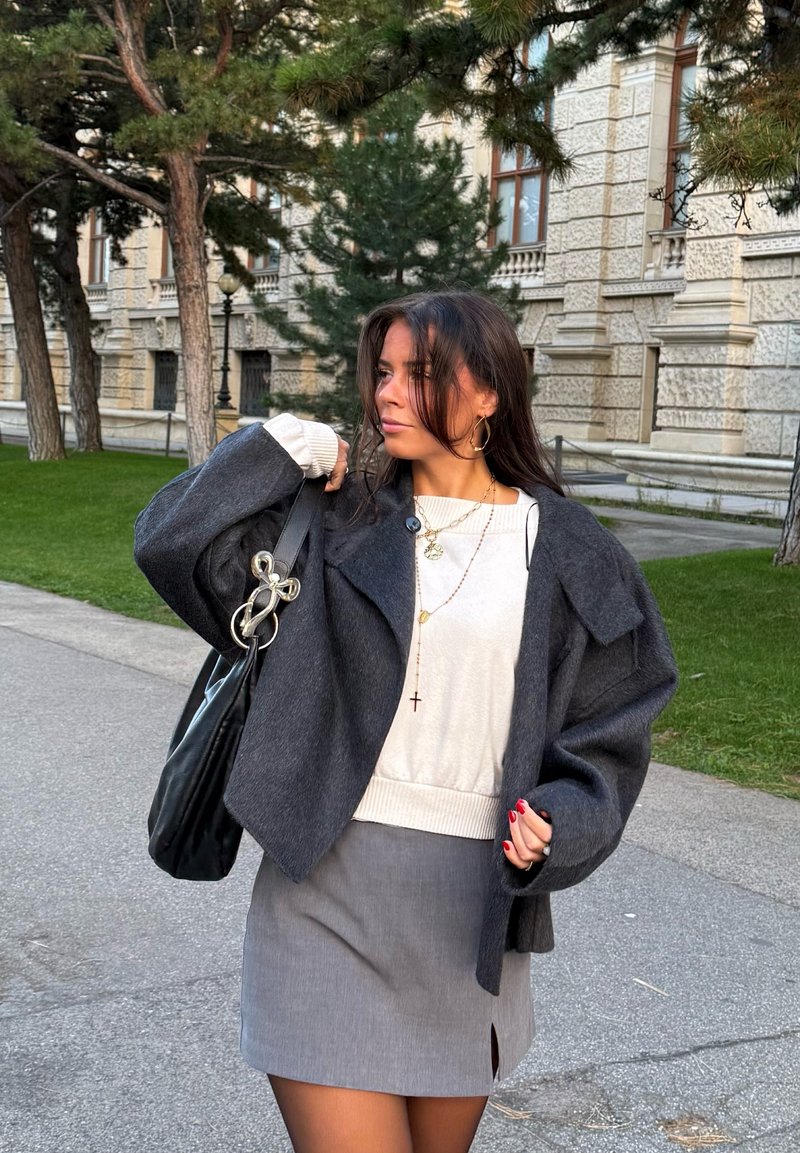 Young woman wearing gray jacket and skirt, white sweater, gold jewelry, and carrying black handbag, standing near trees and ornate building.