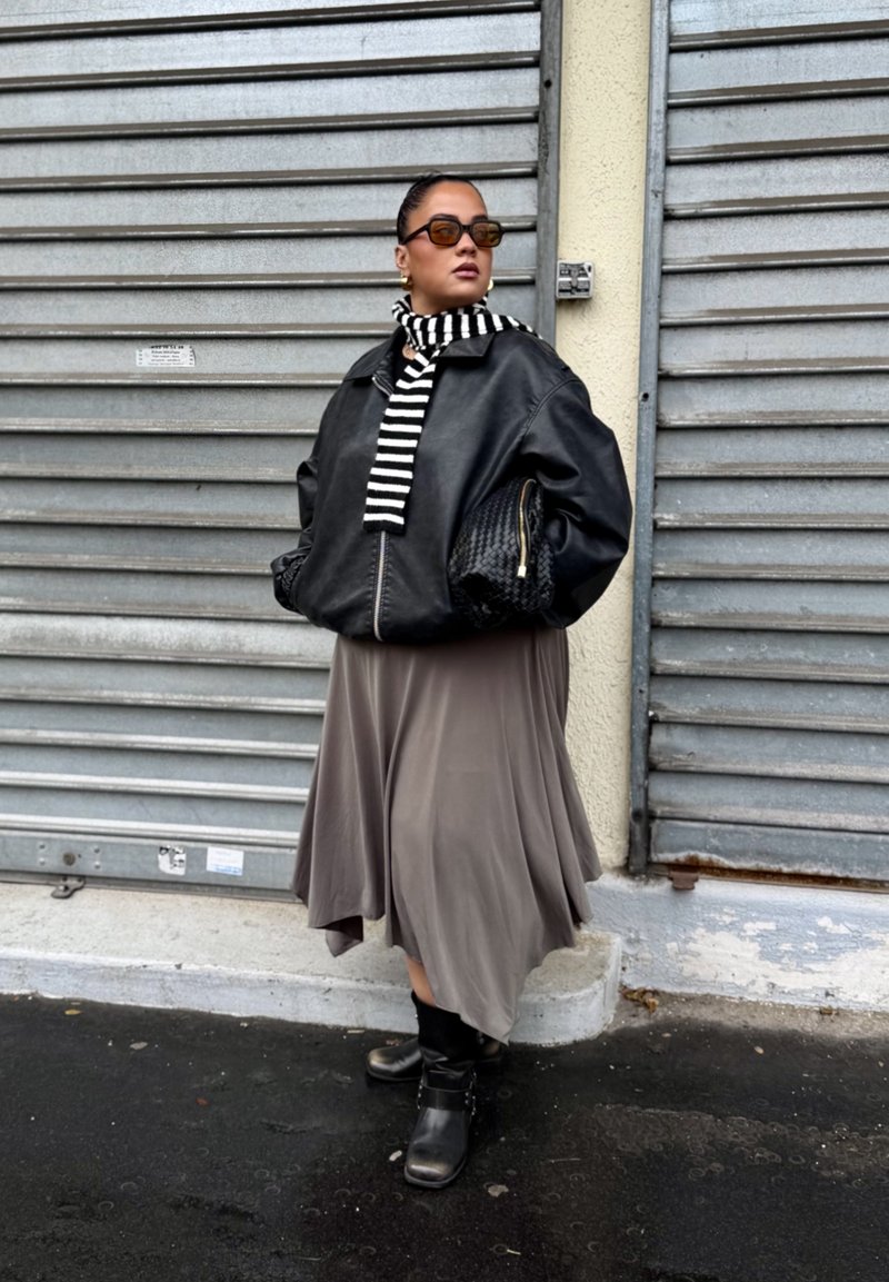 Black leather bomber jacket, striped black-and-white scarf, beige flowy midi skirt, black ankle boots, against a textured gray background.
