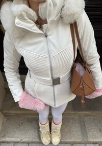 Fehér bélelt dzseki szőrmével bélelt kapucnival, steppelt mintával, övvel a deréknál. Rózsaszín bolyhos kesztyűk, fehér leggings és bézs téli csizma. Barna táska.