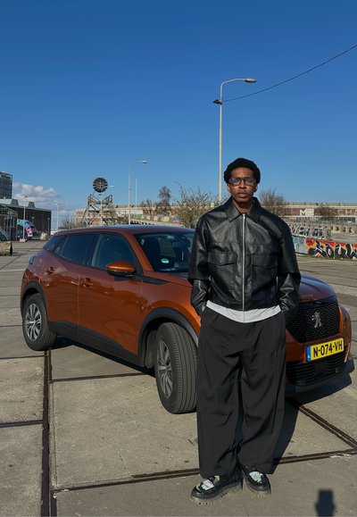 SUV compacto naranja con acentos negros estacionado; una persona con una chaqueta de cuero negra y pantalones anchos negros está de pie junto a él bajo un cielo azul despejado.