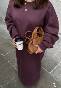 Lange bordeauxrode sweatshirt jurk met oversized mouwen, bruine leren handtas en een koffiekopje met de tekst "aura." op een achtergrond van stenen straat.