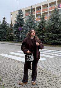 Bruine oversized jas, bijpassende broek en bruine schoenen met patroon. Grijze en zwarte gevlekte tas. Op de achtergrond bomen en een gebouw.