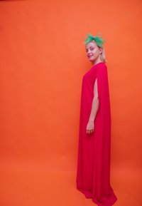 Μακριά, ρέουσα φόρεμα από ζωντανό ροζ ύφασμα, με μανίκια τύπου κάπα. Ένα πράσινο διχτυωτό φασιανί φορεμένο στο κεφάλι εναντίον πορτοκαλί φόντου.