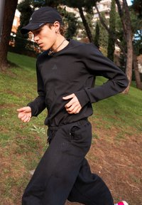 Young person jogging outdoors on grass, wearing a black cap, black long-sleeve top, black trousers, and trainers with pink details.