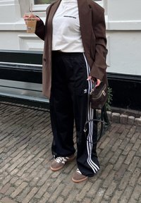Svarte treningsbukser med hvite striper, en hvit t-skjorte med grafikk, en brun oversized blazer, og brune sneakers med hvite detaljer.