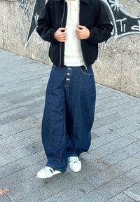 Løse mørkeblå denimjeans med knapgylp, kombineret med en hvid, ribstrikket top og sort jakke med lynlås. Hvide sneakers med mørke detaljer.