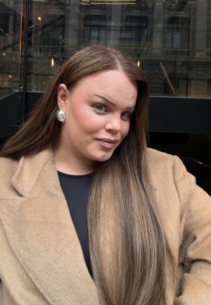 Joven mujer con cabello largo y castaño, que lleva pendientes plateados y un abrigo beige, posando al aire libre en un entorno urbano con reflejos de cristal y edificios.