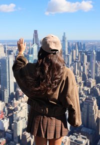 Brun kasket i kordfløjl, oversized brun jakke og et ternet plissert skørt. Langt mørkt hår flyder ned ad ryggen, mens hun vinker mod byens skyline.