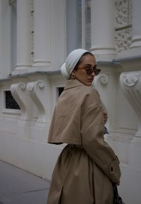 Tan cropped trench coat med knapdetaljer, båret over et hvidt hovedtørklæde. Skildpaddet-brun solbriller og øreringe tilføjer accenter. Glat tekstur.