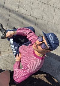 Una persona è seduta su una superficie di pietra, indossando un maglione a righe rosa, pantaloni da jogging blu con strisce bianche e stivali neri. Berretto blu e occhiali da sole.