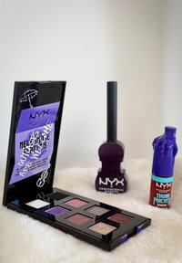 Paleta cieni do oczu NYX Professional Makeup z różnymi odcieniami, fioletowy przedmiot w kształcie gitary oraz fioletowy produkt do ust w kształcie ręki na białej powierzchni.
