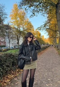 Čierna kožená skrátená bunda, biela košeľa na gombíky, zelená sukňa, čierne pančuchy, čižmy po kolená; mestská jesenná scéna so žltými stromami.