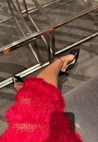 Sapatos de salto alto pretos apontados com acabamento brilhante. Um vestido vermelho com uma bainha texturizada e com franjas, e sobreposição de renda é visível acima do tornozelo.