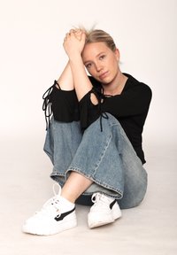 Μαύρη μπλούζα με δεμένα μανίκια, ανοιχτό μπλε παντελόνι σε φαρδιά γραμμή και λευκά sneakers με μαύρες λεπτομέρειες. Απλή, μινιμαλιστική εμφάνιση σε ουδέτερο φόντο.