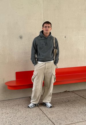 Sudadera gris con logo, pantalones cargo de pana beige y zapatillas plateadas. De pie junto a un banco curvado rojo brillante contra una pared de concreto.